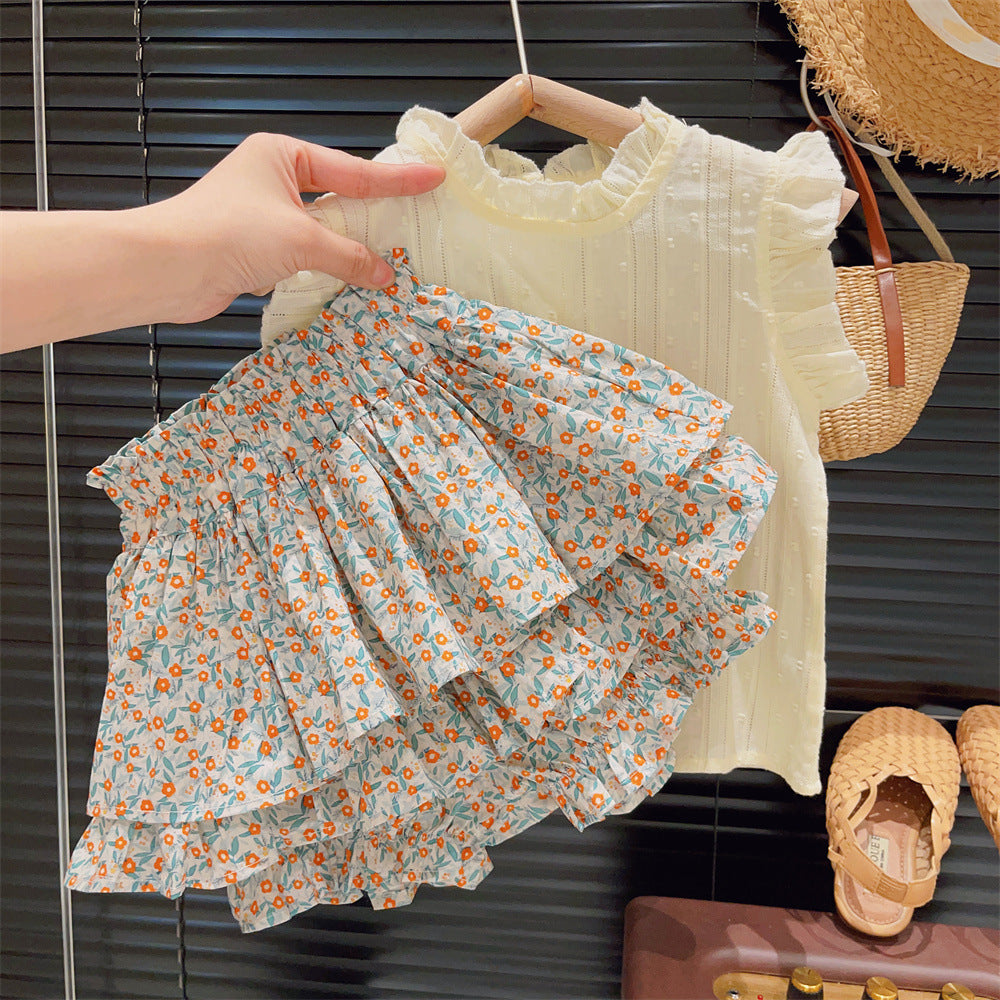 Hand holding a floral tiered skirt in front of a cream ruffled sleeveless blouse on a hanger, with woven bag and sandals nearby