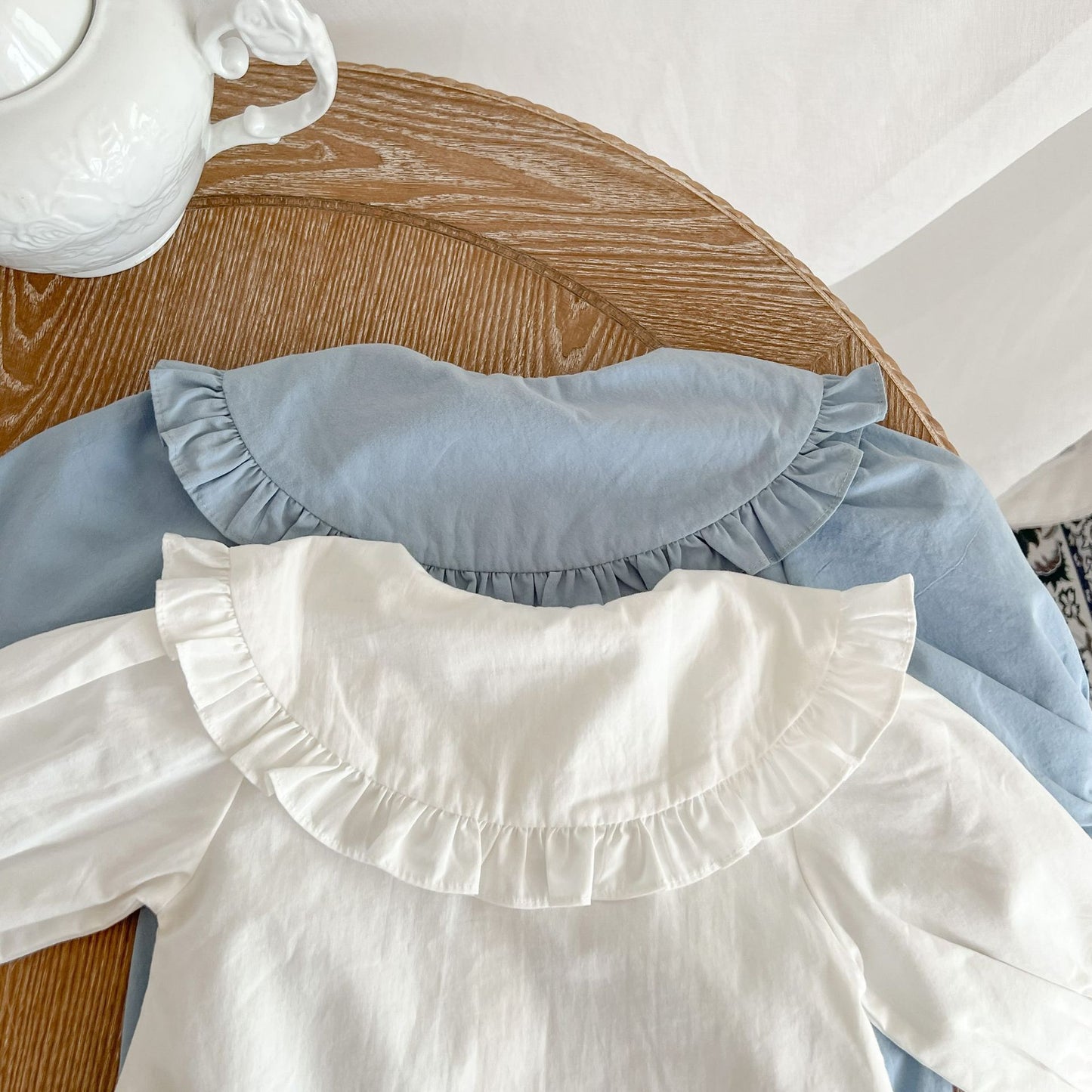 Toddler clothing with ruffled collars in white and light blue on wooden table near white teapot