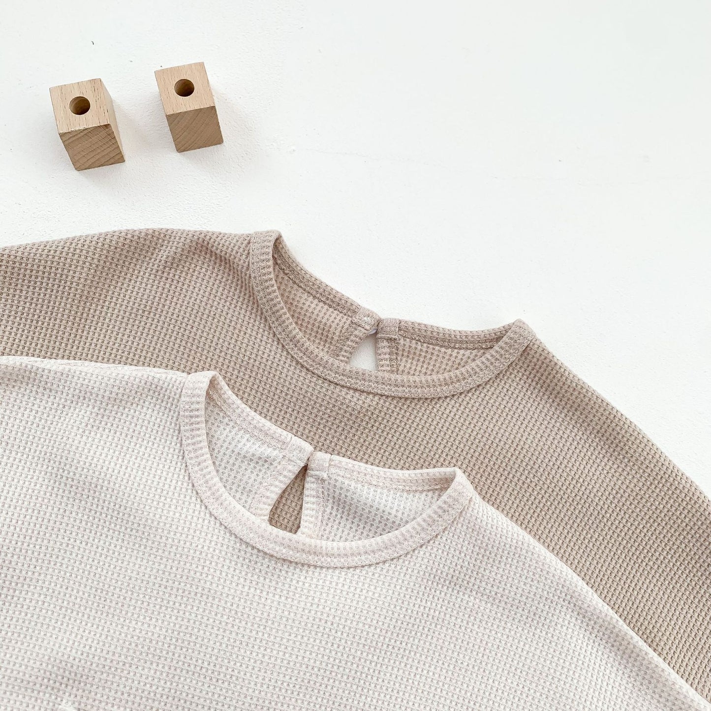 Two toddler waffle knit shirts in beige and cream with round necks laid on white surface with wooden blocks