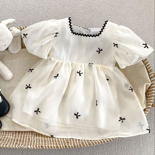 Toddler's cream dress with black bow print, black shoes with white socks, and plush bunny toy in woven basket